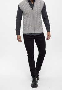 Lysegrå kabelstrikket vest med lynlås, båret over en mørkegrå skjorte, kombineret med sorte slim-fit bukser og sorte snøresko.