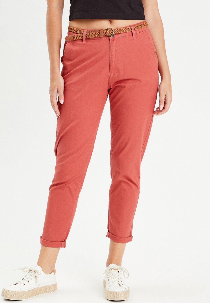 Pantalones de color óxido fabricados en algodón, con dobladillo enrollado, bolsillos laterales y un cinturón trenzado marrón. Combinados con zapatos blancos.