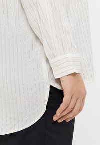 Chemise blanche à rayures avec de fines lignes métalliques dorées, manches longues et poignets à boutons. Le tissu semble léger et texturé.