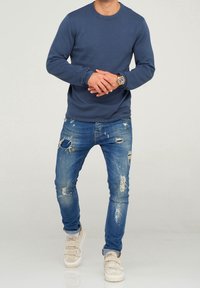 Marineblaues Langarm-Crewneck-Sweatshirt, ausgefranste blaue Jeans mit Rissen und beiges Sneakers mit Klettverschlüssen. Uhr am Handgelenk.