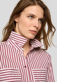 Red and white striped shirt with a high collar, front pocket, and button closure, featuring a smooth, woven texture.