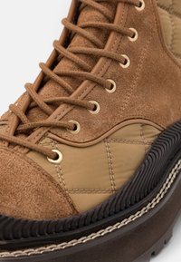 Botte marron à lacets avec des panneaux en daim et en tissu, des œillets métalliques, des détails cousus, et une semelle extérieure épaisse en caoutchouc noir avec un motif antidérapant.