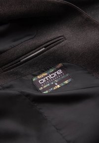 Interior de una prenda de tela de tweed marrón con forro de satén negro y una etiqueta con acentos en verde y negro que dice "ombre QUINTESSENCE M".
