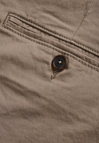 Beige katoenen stof met een gladde textuur, voorzien van een gestikte zak en een ronde bruine knop met ingekerfde tekst.