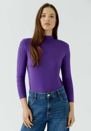 Jeune femme aux longs cheveux bruns porte un haut violet à manches longues et un jean bleu taille haute, debout devant un fond clair uni.