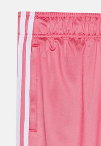 Różowe sportowe krótkie spodenki o gładkiej fakturze, z białymi paskami po bokach i elastycznym paskiem w talii. Z boku widoczna jedna kieszeń.