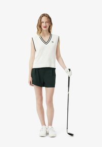 Gilet da golf bianco senza maniche con motivo a rombi e bordi neri, abbinato a pantaloni corti verde scuro. Scarpe da golf bianche e una mazza da golf in mano.
