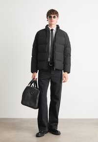Jachetă puffer neagră cu un design matlasat orizontal, purtată peste o cămașă albă și o cravată neagră. Blugi închiși la culoare și o geantă din piele neagră completează look-ul.