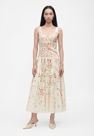 Kvinde iført en ærmeløs, cremefarvet midi-kjole med lyserøde blomsterprint og beige hælede mules, stående foran en ensfarvet hvid baggrund.