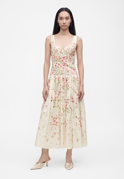 Kvinde iført en ærmeløs, cremefarvet midi-kjole med lyserøde blomsterprint og beige hælede mules, stående foran en ensfarvet hvid baggrund.