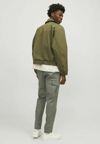 Chaqueta bomber verde oliva con dobladillo elástico, combinada con pantalones cargo gris claro. Zapatillas blancas completan el conjunto. Diseño simple y utilitario.