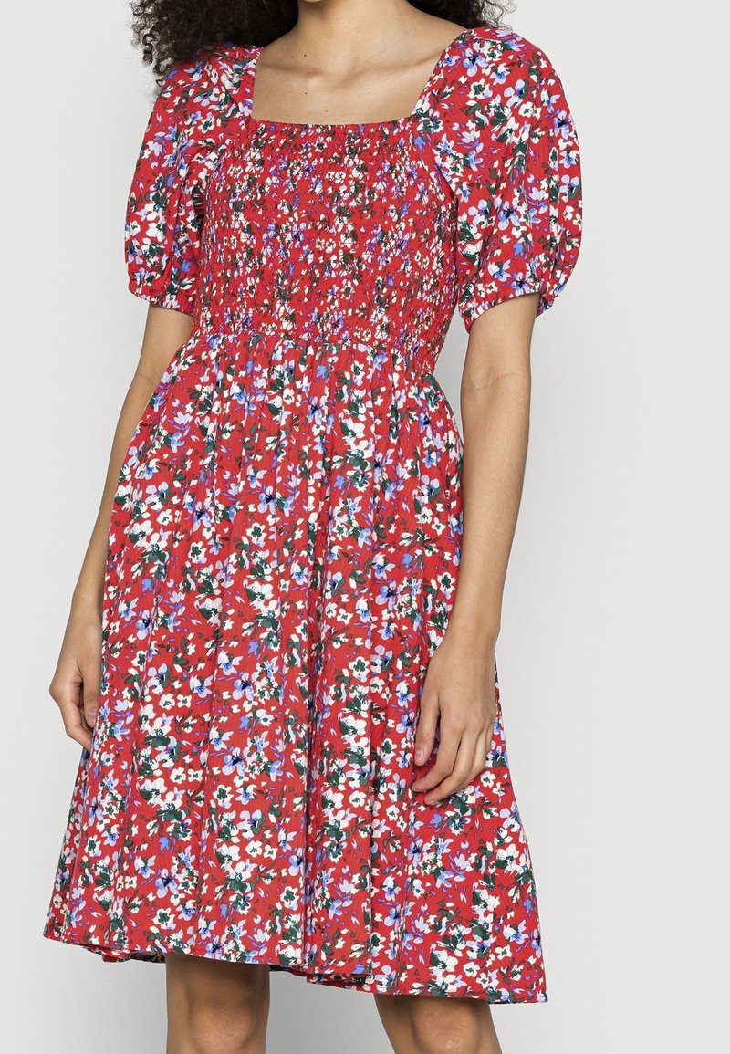 Robe florale rouge avec des manches ballon, un décolleté carré et un corsage froncé. Présente un motif de fleurs blanches, bleues et vertes sur un tissu texturé.