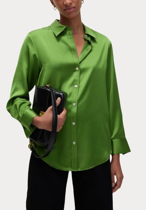 Kobieta ubrana w zieloną satynową koszulę na guziki, trzymająca czarną skórzaną torebkę z złotym zapięciem, na jednolitym tle.