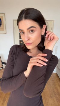 Jeune femme aux cheveux foncés portant un haut marron à manches longues, posant à l'intérieur avec les bras croisés et une expression neutre.