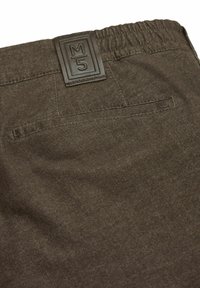 Detailansicht brauner Stoffhosen mit Gesäßtasche, elastischem Bund und einem Lederpatch mit dem "M/5"-Logo.