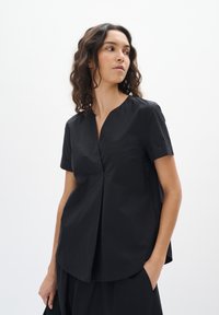 Blusa nera a maniche corte con scollo a V, caratterizzata da un sottile patta e dettagli di piega, realizzata in un tessuto leggero con una texture liscia.