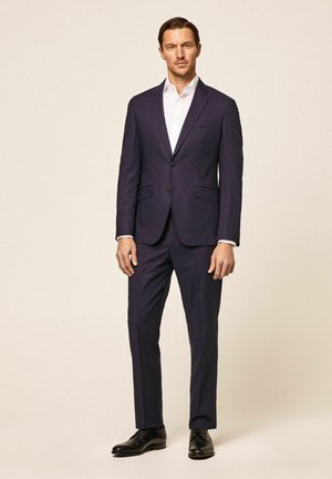 Granatowy garnitur z pojedynczym zapięciem na guzik, klapami ze ścięciem i bocznymi kieszeniami. W zestawie z białą koszulą i czarnymi butami formalnymi.