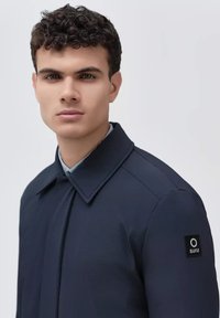 Cappotto navy con colletto classico, realizzato in un tessuto strutturato e con una patch brandizzata sulla manica sinistra. Design semplice con una vestibilità su misura.