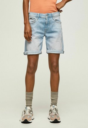 Person iført lyseblå op rullede jeansshorts, beige sokker og flerfarvede sneakers står mod en ensfarvet baggrund.
