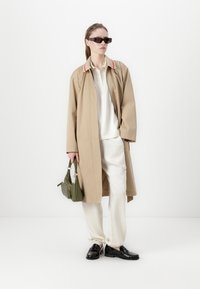 Beige pitkä takki, jossa on punaiset kaulukset, valkoinen poolopaita, kermaiset housut, mustat loafers-kengät ja pieni vihreä käsilaukku. Sileä rakenne ja moderni muotoilu.