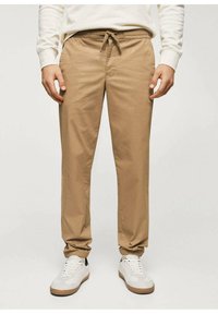 Pantalon beige léger avec taille élastique et cordon de serrage, présentant une texture lisse. Associé à des baskets blanches et un sweat crème.
