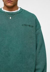Teal groene sweatshirt van zacht materiaal met een ontspannen pasvorm, voorzien van ribgebreide boorden en hals. Inclusief geborduurde tekst op de borst.