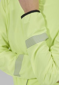 Lehká bundu neonově žluté barvy s reflexními šedými tečkovanými akcenty na rukávech a černým elastickým manžetovým zakončením. Hladká textura a přiléhavý design.