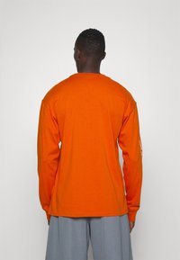 Long-sleeve orange t-shirt with a crew neckline and textured fabric. It features a logo printed on one sleeve and relaxed fit.
