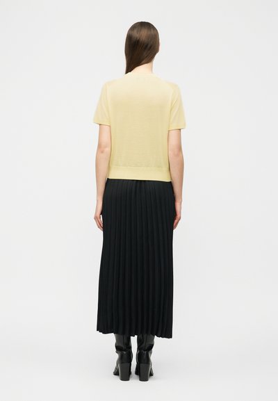 Γυναίκα με ανοιχτό κίτρινο τοπ με κοντά μανίκια, μαύρη πλισέ μίντι φούστα και μαύρα μποτάκια με τακούνι, στέκεται γυρισμένη με την πλάτη σε ένα απλό φόντο.