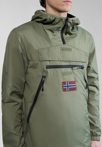Chaqueta ligera verde oliva con capucha, que cuenta con un bolsillo en el pecho con cremallera, acentos negros y un parche de la bandera noruega.