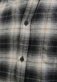 Camisa de franela a cuadros con tonos negros, blancos y grises, con acentos marrones. Dos botones negros y un detalle de bolsillo en el pecho. Textura suave.