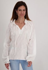 Witte blouse van lichtgewicht stof met zigzagpatroon en opengewerkte details; heeft een geplooide halslijn en elastische manchetten.