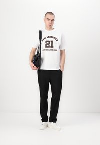 Hvit t-skjorte med brun "21" og "Karl Lagerfeld" tekst, kombinert med svarte slim-fit bukser og hvite sneakers. Liten svart bag med stropp.