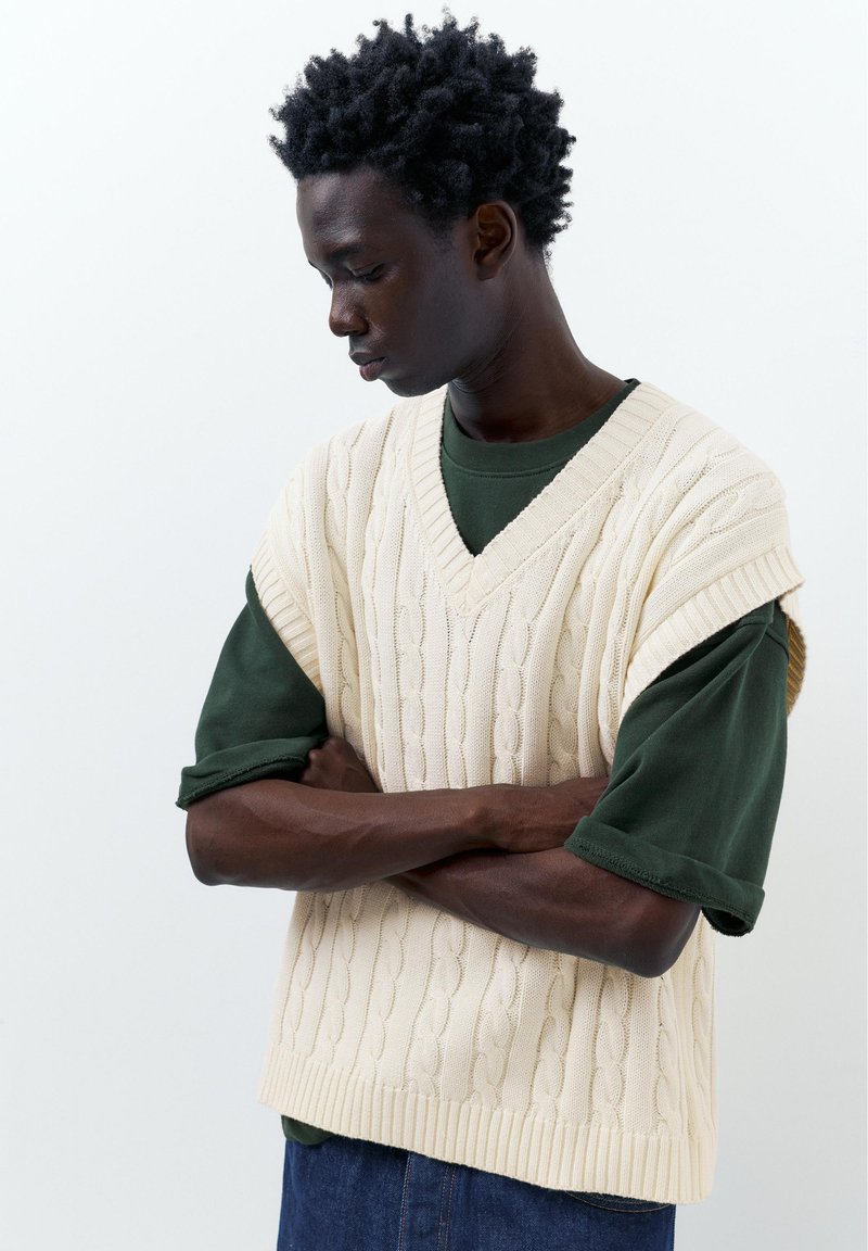 Jeune homme aux bras croisés regarde vers le bas, portant une chemise vert foncé sous un pull sans manches en maille torsadée crème.
