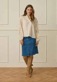 Femme portant un chemisier crème à manches en dentelle, une jupe en jean bleue et des bottes hautes couleur tan, debout sur un sol en bois devant un mur beige à panneaux.