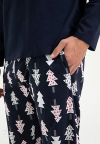 Męski zestaw piżamowy z granatową bluzą i wzorzystymi spodniami w białe drzewa, czerwone kropki i szare akcenty na ciemnym tle.