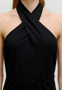 Schwarzes Trägerkleid mit einem Kreuzkragen, das aus einem strukturierten Stoff besteht. Das Design hat eine A-Linie-Form ohne sichtbare Verschlüsse.