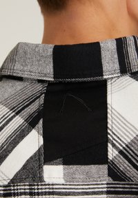 Gros plan sur le cou d'une personne et le col d'une chemise à carreaux noir et blanc avec un écusson en tissu noir dans le dos.