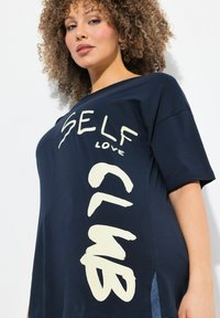 Camiseta de color azul marino con mangas cortas, que presenta un gran texto blanco "SELF LOVE CLUB" en varios estilos. Un detalle con abertura lateral añade interés.