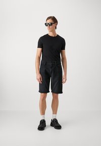 Camiseta negra de manga corta, pantalones cortos de denim oscuro con dobladillos enrollados y zapatillas negras. El modelo lleva gafas de sol; fondo liso.