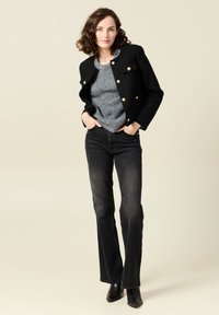 Giacca nera corta con bottoni dorati, maglione grigio e jeans neri a campana. Modella con stivali neri alla caviglia, su uno sfondo beige chiaro.