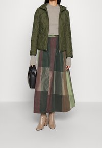 Quilted olivengrøn jakke, lys beige ribstrikket sweater, multikolor maxi nederdel med geometriske mønstre, beige hæl støvler, sort håndtaske.