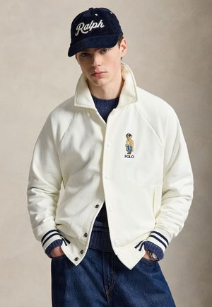 Млад мъж, носещ бяло яке Polo с бродирана мечка, син пуловер, дънкови панталони и тъмносиня шапка с надпис "Ralph".