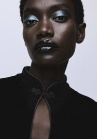 Prenda negra de cuello alto con una abertura en forma de llave, que presenta intrincados cierres florales. El modelo lleva un maquillaje audaz en tonos azul y negro.