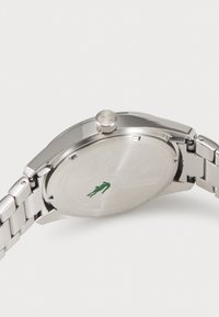 Ceas din oțel inoxidabil argintiu, cu un carcasă netedă, coronă și un logo de crocodil verde pe spate. Dispune de o brățară din metal periat.