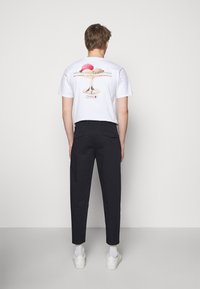 Witte t-shirt met kleurrijke ijsje graphic op de achterkant, gecombineerd met donkere broek en witte sneakers; model kijkt van de camera weg.