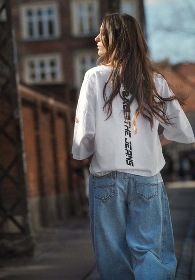 Mlada žena s dugom kosom u oversized bijeloj majici s crnim natpisom i širokim plavim trapericama hoda na otvorenom u urbanom okruženju.