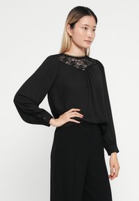 Svart bluse med lange ermer, blonde detaljer ved halsen, plisserte foran og knapping ved mansjettene. Glatt tekstur og avslappet passform.