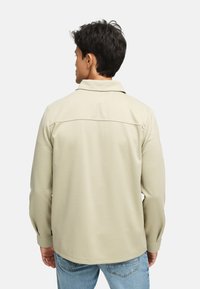 Lys beige jakke med krage, lange ermer og en jevn tekstur. Har en yoke-detalj på baksiden og er kombinert med lyseblå jeans.
