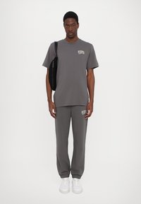 Grijze katoenen T-shirt met logo op de borst, gecombineerd met bijpassende grijze joggers. Model draagt witte sneakers en houdt een zwarte schoudertas vast.